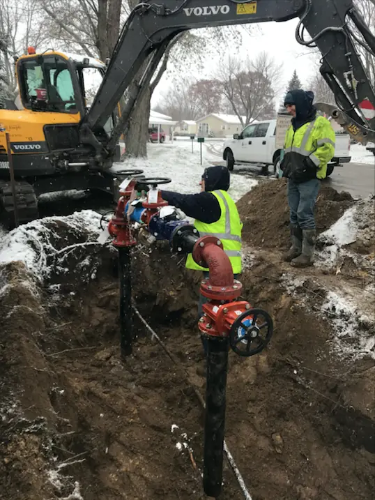 A two-person City Plumbing crew in hi-vis vests installing a commercial water line and valve in an open trench